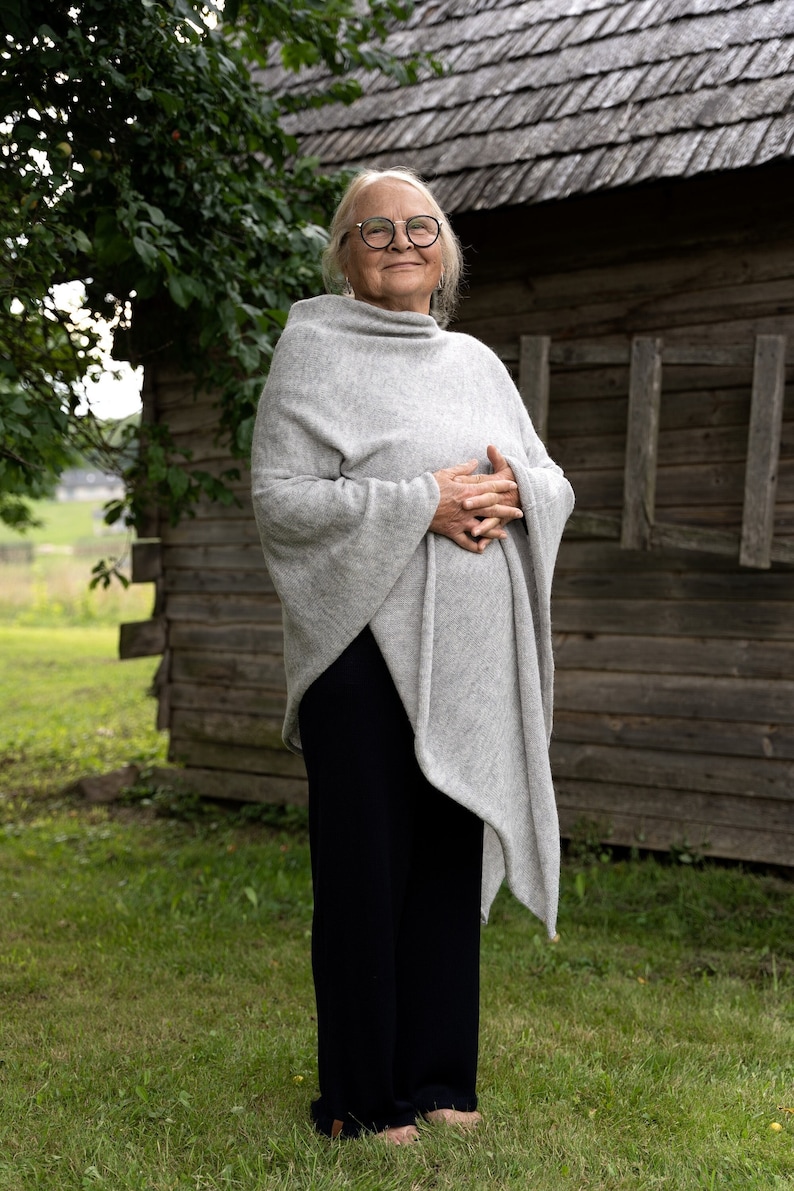 May include: A woman wearing a grey poncho and black trousers stands in front of a wooden building. She is smiling and looking at the camera.