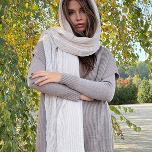 May include: A white knitted scarf with a ribbed texture. The scarf is draped around the neck of a person wearing a brown knitted sweater.