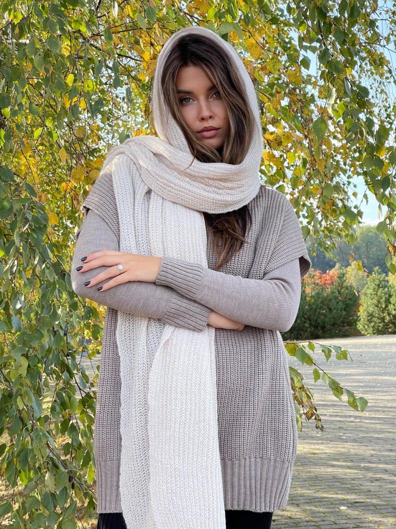 May include: A white knitted scarf with a ribbed texture. The scarf is draped around the neck of a person wearing a brown knitted sweater.