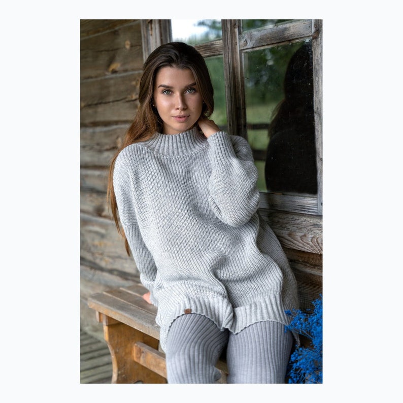 K&ouml;nnte beinhalten: Ein grauer Strickpullover mit hohem Kragen und langen &Auml;rmeln. Der Pullover ist locker geschnitten und hat eine gerippte Struktur. Das Model tr&auml;gt den Pullover mit grauen Leggings.