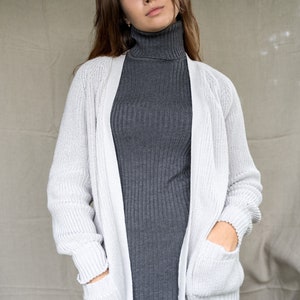May include: A white knitted cardigan with pockets, worn over a gray turtleneck sweater.