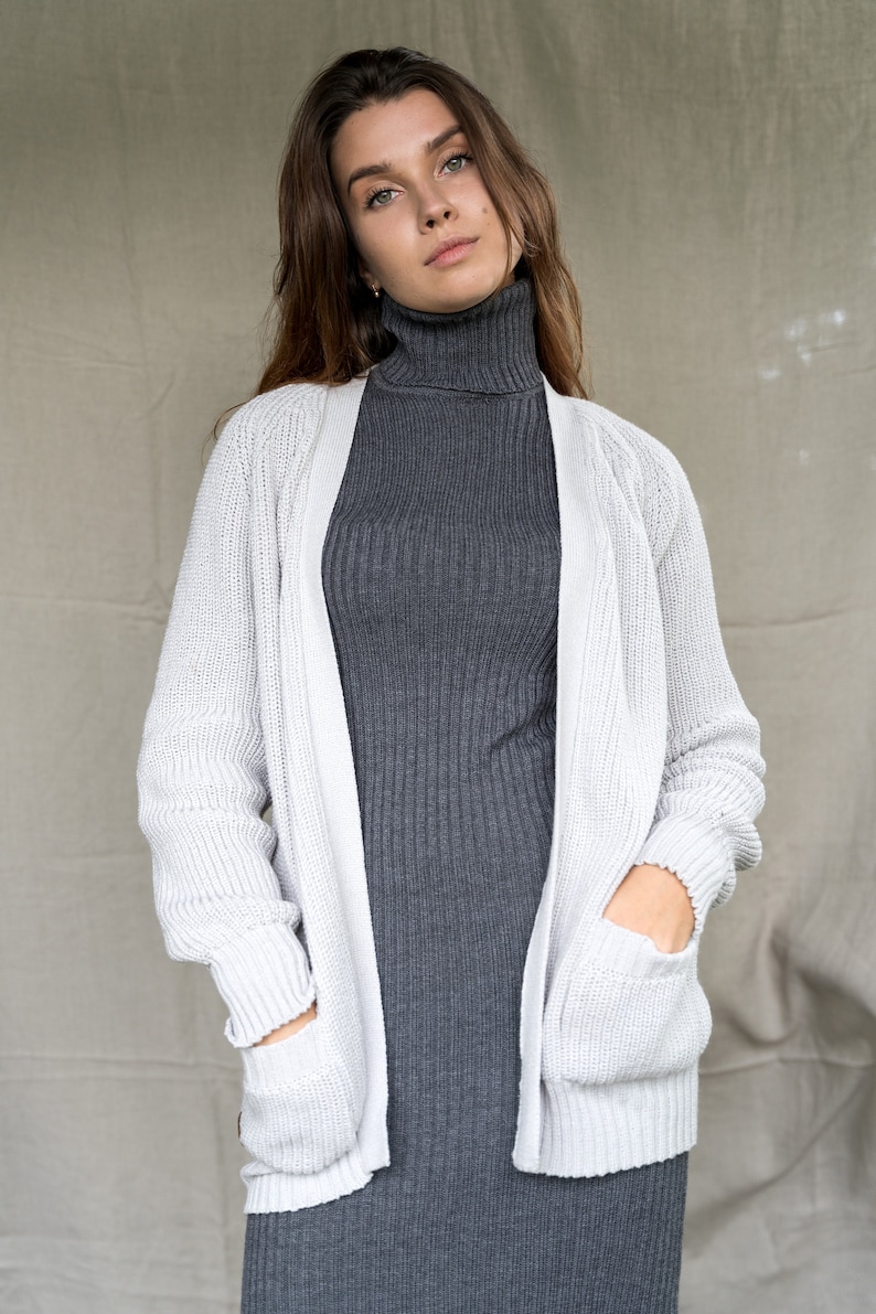 May include: A white knitted cardigan with pockets, worn over a gray turtleneck sweater.