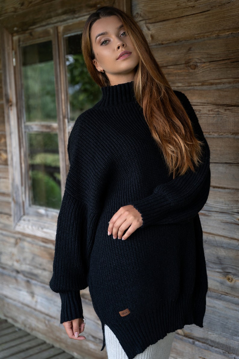K&ouml;nnte beinhalten: Ein schwarzer, &uuml;bergro&szlig;er Strickpullover mit Stehkragen und langen &Auml;rmeln. Der Pullover hat eine gerippte Struktur und ein kleines braunes Etikett mit dem Wort "MADE" darauf.