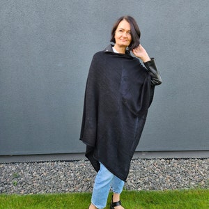 May include: A woman wearing a black poncho with leather sleeves and blue jeans. She is standing in front of a grey wall and green grass.