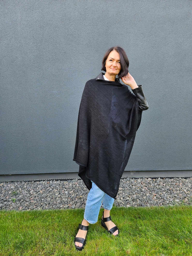 May include: A woman wearing a black poncho with leather sleeves and blue jeans. She is standing in front of a grey wall and green grass.