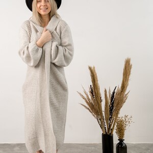 May include: A woman wearing a long, light gray knitted cardigan, a black fedora hat, and brown leather sandals. She is standing in front of a white wall with dried flowers in black vases.