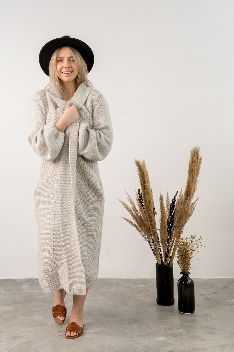 May include: A woman wearing a long, light gray knitted cardigan, a black fedora hat, and brown leather sandals. She is standing in front of a white wall with dried flowers in black vases.