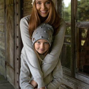 May include: Two people wearing grey knitted hats with pom poms. The hats have a textured pattern. The person in the front is a child and the person in the back is an adult. They are both smiling and looking at the camera.