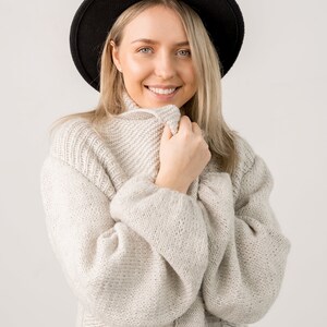 May include: A woman wearing a white knitted sweater with a black fedora hat. She is smiling and looking at the camera.