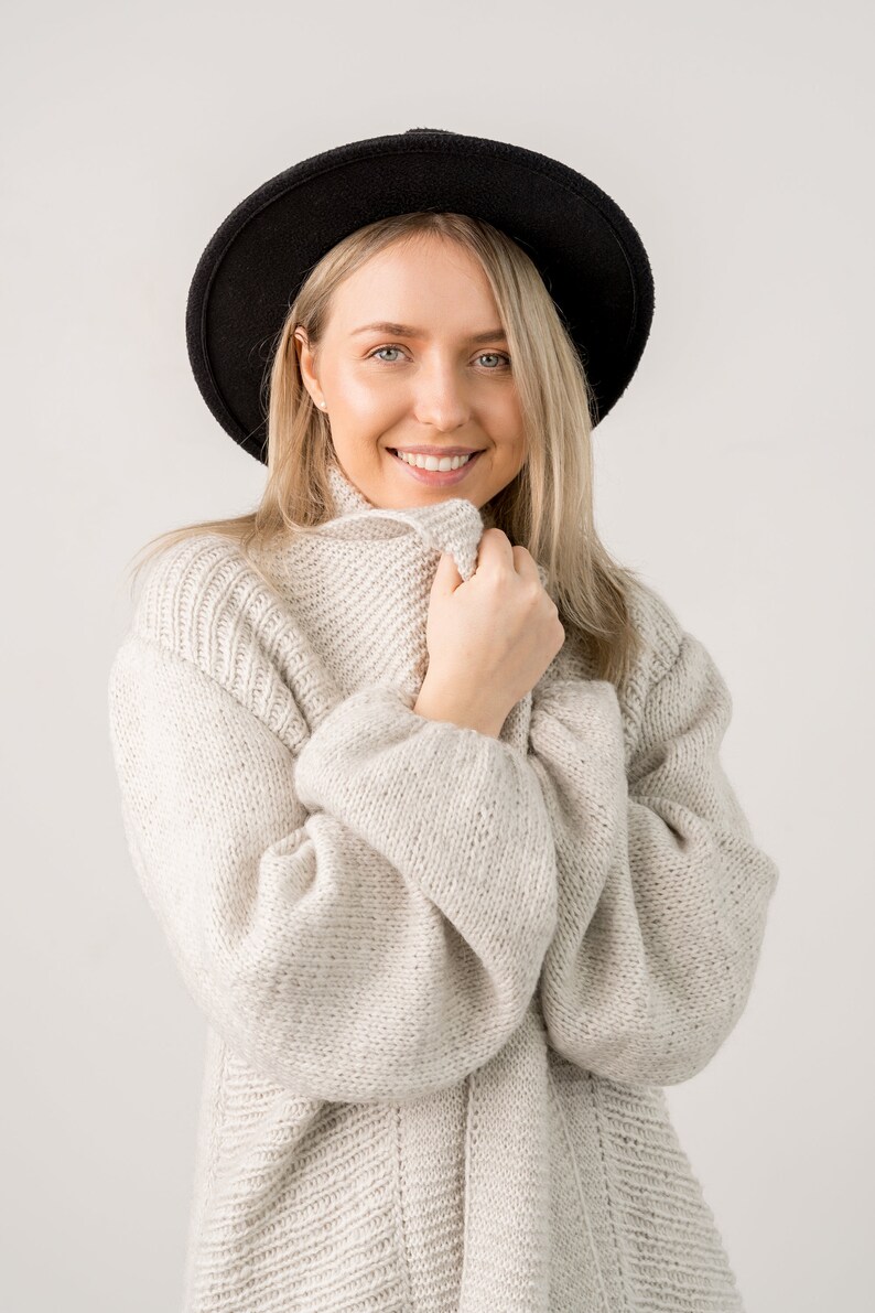 May include: A woman wearing a white knitted sweater with a black fedora hat. She is smiling and looking at the camera.