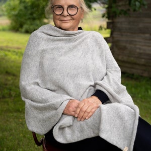 May include: A woman wearing round glasses sits on a red wooden chair with a grey knitted shawl draped around her shoulders. She is smiling and looking at the camera.