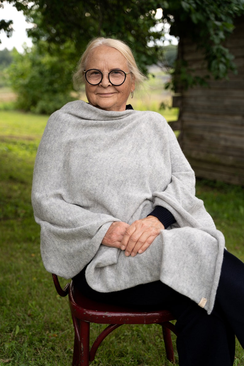 May include: A woman wearing round glasses sits on a red wooden chair with a grey knitted shawl draped around her shoulders. She is smiling and looking at the camera.