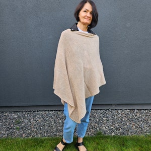 May include: A woman wearing a beige poncho with a black leather collar and blue jeans. She is standing in front of a grey wall and green grass.