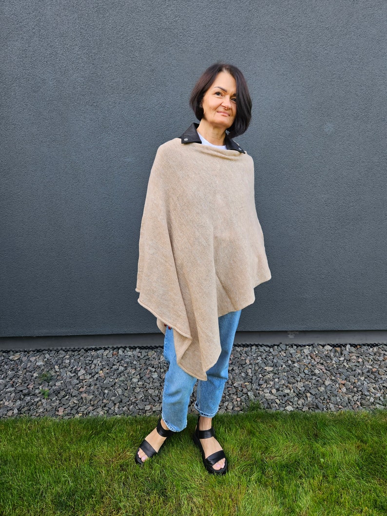 May include: A woman wearing a beige poncho with a black leather collar and blue jeans. She is standing in front of a grey wall and green grass.