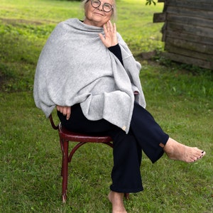 May include: An older woman wearing glasses sits on a red chair in a grassy field. She is wearing a grey shawl and dark trousers. Her feet are bare.