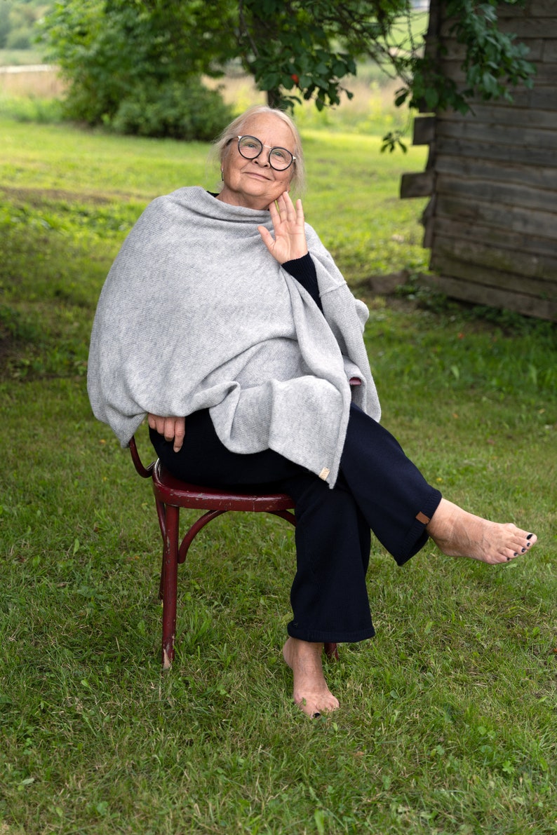 May include: An older woman wearing glasses sits on a red chair in a grassy field. She is wearing a grey shawl and dark trousers. Her feet are bare.