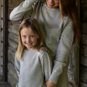 May include: A woman and a girl wearing matching gray sweaters. The woman is smiling and looking at the camera. The girl is smiling and looking at the camera. They are standing in front of a wooden wall.