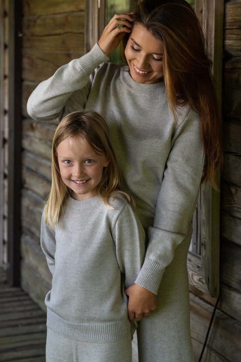 May include: A woman and a girl wearing matching gray sweaters. The woman is smiling and looking at the camera. The girl is smiling and looking at the camera. They are standing in front of a wooden wall.