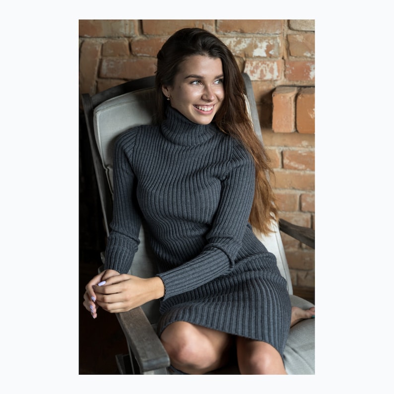 May include: A woman wearing a gray ribbed turtleneck sweater dress, sitting in a chair with a brick wall in the background.