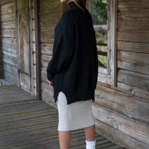 K&ouml;nnte beinhalten: Eine Frau in einem schwarzen Pullover und einem beigefarbenen Rock, die auf einer Holzveranda steht. Sie tr&auml;gt wei&szlig;e Socken und braune Stiefel.