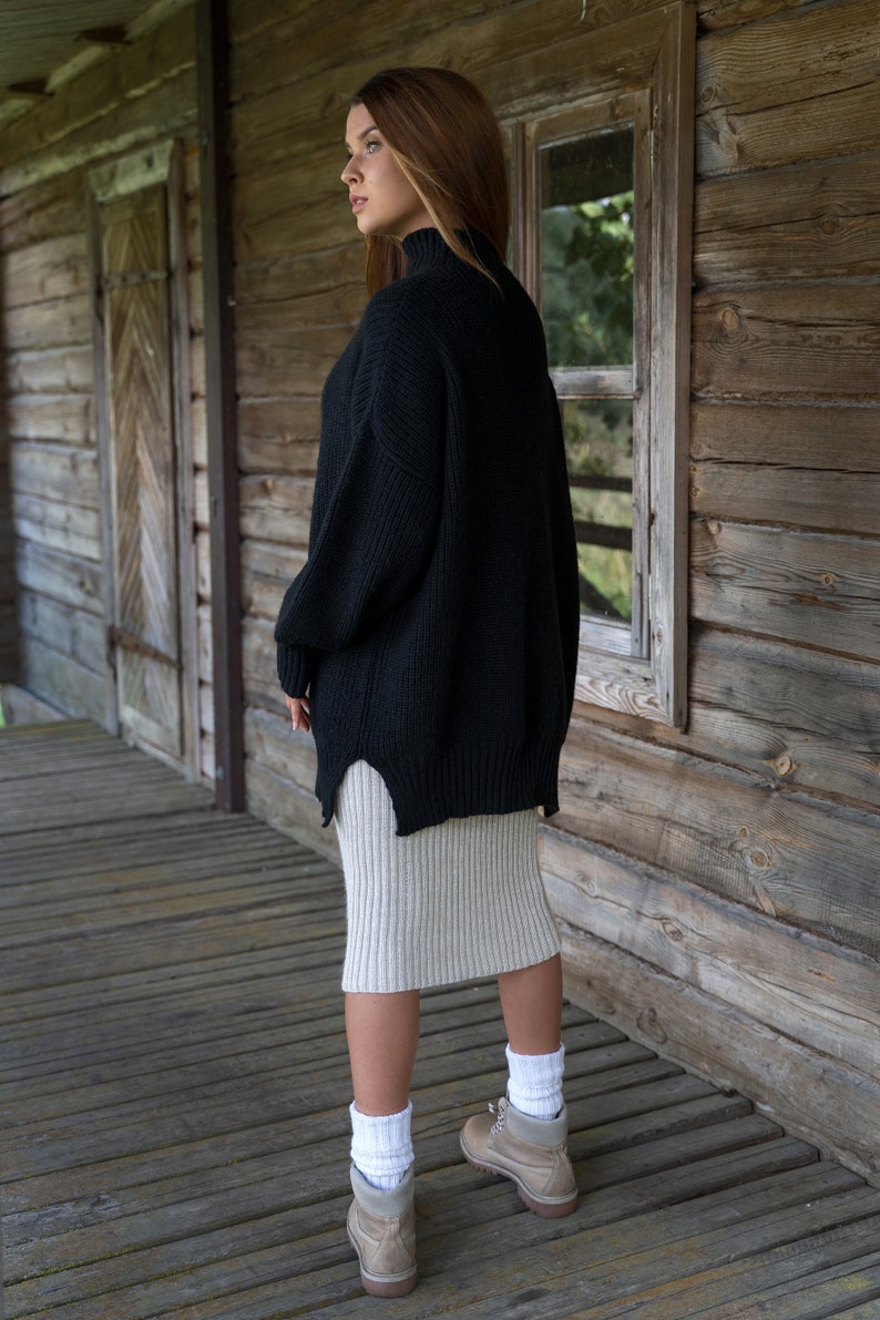 K&ouml;nnte beinhalten: Eine Frau in einem schwarzen Pullover und einem beigefarbenen Rock, die auf einer Holzveranda steht. Sie tr&auml;gt wei&szlig;e Socken und braune Stiefel.