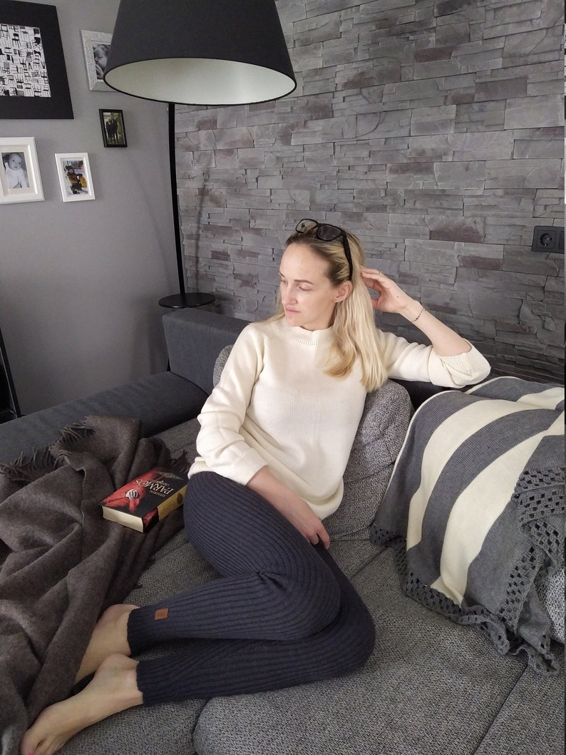 May include: A woman wearing a white sweater and dark gray ribbed leggings sits on a gray couch. She is holding a book titled "Pride and Prejudice" in her hands. The couch has a gray and white striped throw pillow and a brown throw blanket.