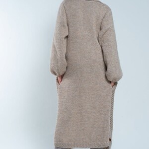 May include: A long, beige knitted cardigan with a high collar and long sleeves. The cardigan is made of a soft, chunky yarn and has a relaxed fit.