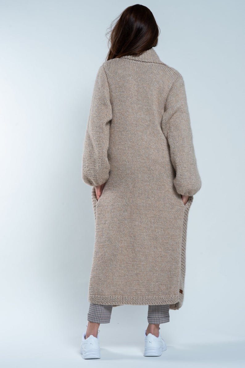 May include: A long, beige knitted cardigan with a high collar and long sleeves. The cardigan is made of a soft, chunky yarn and has a relaxed fit.