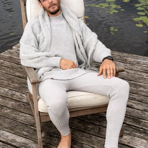 May include: A person wearing a gray knitted sweater and matching pants sits in a wooden chair on a wooden dock. The person is barefoot and has a watch on their wrist. The dock is over a body of water with lily pads.