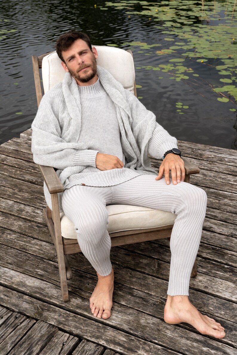 May include: A person wearing a gray knitted sweater and matching pants sits in a wooden chair on a wooden dock. The person is barefoot and has a watch on their wrist. The dock is over a body of water with lily pads.