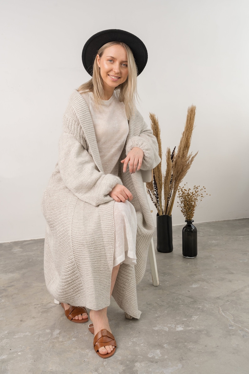 May include: A woman wearing a light gray knitted cardigan, a white dress, and brown sandals. She is sitting on a white stool and wearing a black hat. There are dried flowers in vases behind her.
