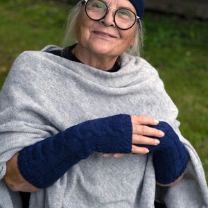 May include: A woman wearing a grey knitted poncho and blue knitted fingerless gloves. She is wearing a blue headband and glasses.