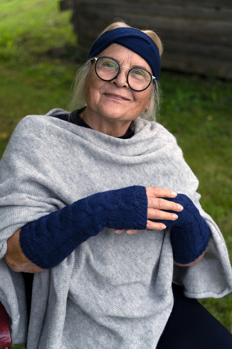 May include: A woman wearing a grey knitted poncho and blue knitted fingerless gloves. She is wearing a blue headband and glasses.