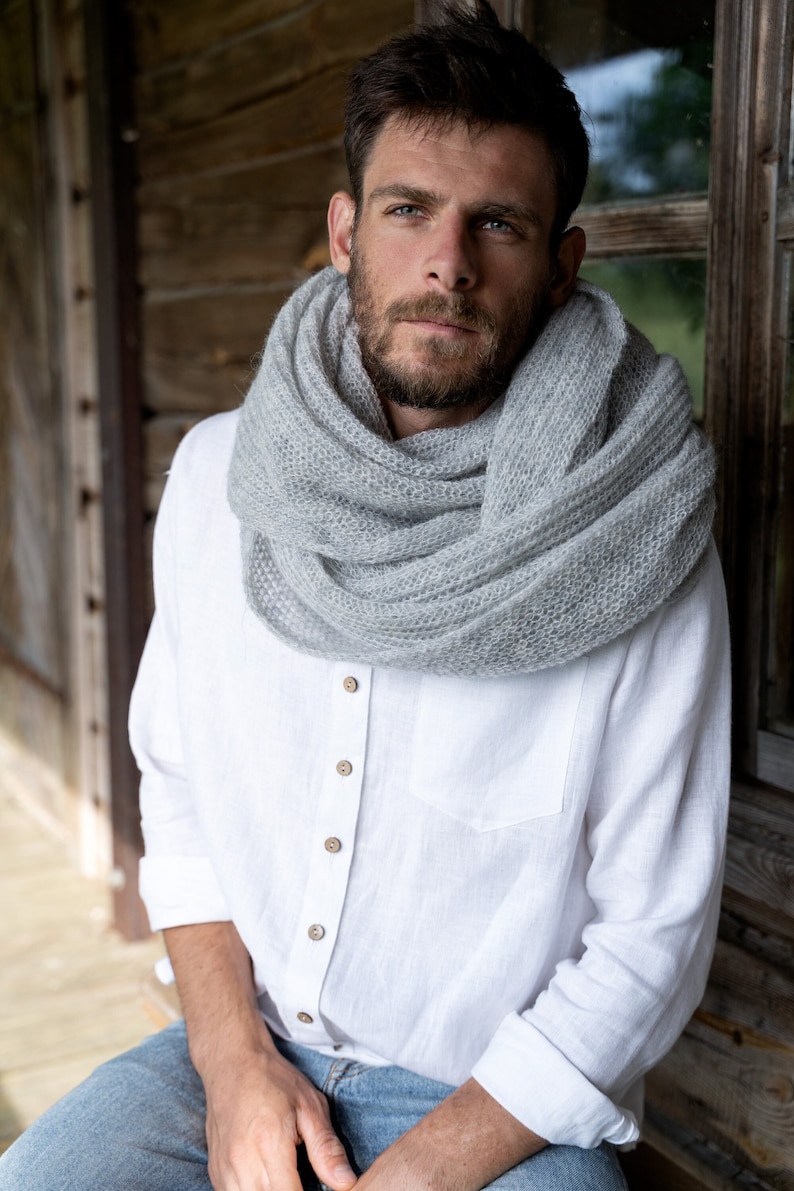 May include: A man wearing a white linen shirt with wooden buttons and a gray knitted scarf. He is sitting on a wooden porch.