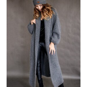 May include: A long, gray knitted cardigan with a shawl collar and long sleeves. The cardigan is made of a soft, fuzzy yarn and has a relaxed fit.