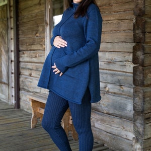 May include: A pregnant person wearing a dark blue knitted sweater and matching leggings. The person is standing in front of a wooden wall and smiling at the camera.