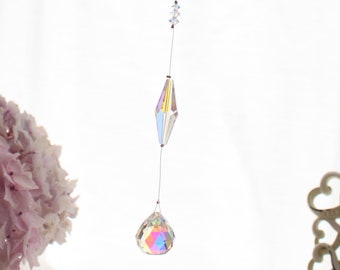 Op de afbeelding: Een kristallen zonenvanger met iriserende regenboogkleuren hangt aan een wit plafond. De zonenvanger heeft een traanvormig kristal aan de onderkant en twee ruitvormige kristallen erboven.