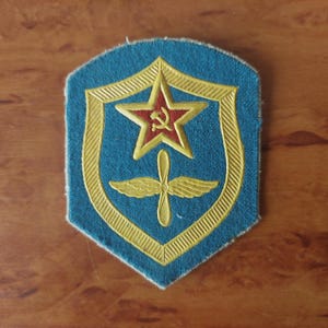 May include: A vintage patch with a blue background and a gold shield design. Inside the shield is a red star with a hammer and sickle, and a gold propeller. The patch is likely from the Soviet era.