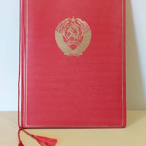 May include: A red book with a gold emblem featuring a hammer and sickle, wheat, and a star. The book has a gold border and a red tassel bookmark. The cover has a textured appearance.