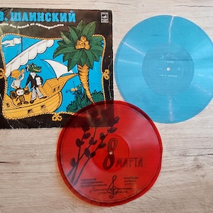Könnte beinhalten: Sammlung von Vintage-Schallplatten. Das Albumcover zeigt eine Cartoon-Illustration eines Bootes mit Figuren und Text auf Russisch. Eine hellblaue Schallplatte und eine transparente rote Schallplatte mit Text sind ebenfalls sichtbar.
