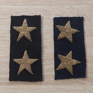 May include: Two dark blue fabric patches, each featuring two gold-coloured, embroidered stars. The stars have a detailed, spiral design. The patches are rectangular and are likely uniform insignia.