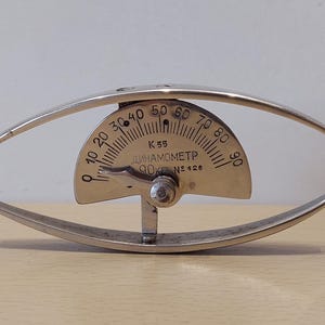 Könnte beinhalten: Ein silbernes Vintage-Dynamometer mit einem gebogenen, ovalen Rahmen. Das Gerät verfügt über eine halbkreisförmige Anzeige mit Markierungen von 0 bis 90, und der Text "ДИНАМОМЕТР" ist sichtbar. Das Instrument dient zur Messung der Kraft.