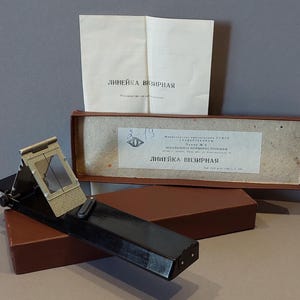 Può includere: Un dispositivo di mira nero e argento vintage con un mirino incernierato, appoggiato su una scatola marrone. Il dispositivo è accompagnato da una scatola di cartone marrone e da un foglio di carta bianca piegato con testo cirillico. Il testo sulla scatola e sulla carta è in cirillico.