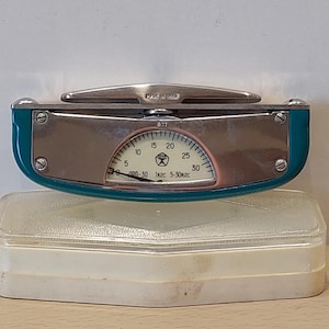 Könnte beinhalten: Eine Vintage-Waage in Türkis und Silber mit einer halbkreisförmigen Anzeige, die Zahlen von 0 bis 30 anzeigt. Die Waage hat einen silbernen Griff und ist mit "Made in USSR" gekennzeichnet. Sie steht auf einem transparenten Kunststoffgehäuse.