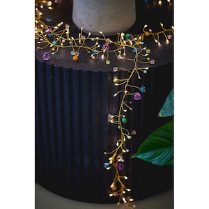 May include: A gold wire garland with small, warm white LED lights and colourful beads in purple, blue, green, and orange. The garland is draped over a dark, textured surface. The beads are in various sizes.