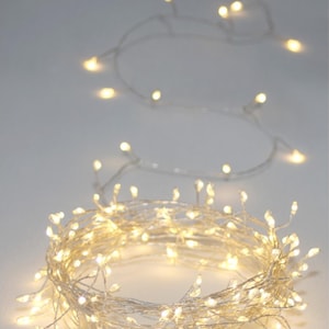 May include: A close-up of a string of warm white LED fairy lights. The lights are arranged in a circular pattern, with some strands extending outwards. The lights are on a thin, clear wire, and the background is a soft white.