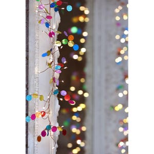 May include: A decorative string of lights featuring colourful, sequined, circular accents. The lights are strung on a thin, brown wire and emit a soft, warm glow. The sequins are in various colours, including blue, pink, green, and gold. The background is blurred with additional lights.