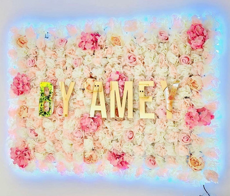 May include: A white and pink flower wall with the words "BY AMEY" in gold letters.