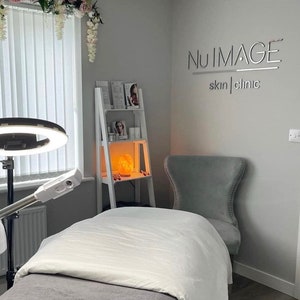 May include: A white treatment table with a white sheet and a gray blanket. A ring light is on a stand to the left of the table. A white shelf with products and a salt lamp is in the background. A gray chair is in the corner of the room. The text "NU IMAGE skin | clinic" is on the wall.