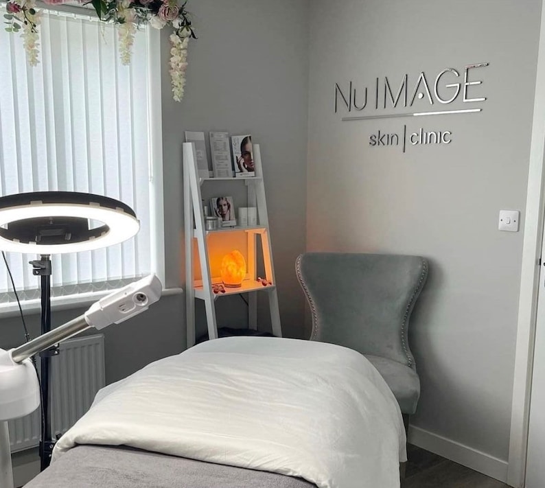 May include: A white treatment table with a white sheet and a gray blanket. A ring light is on a stand to the left of the table. A white shelf with products and a salt lamp is in the background. A gray chair is in the corner of the room. The text "NU IMAGE skin | clinic" is on the wall.